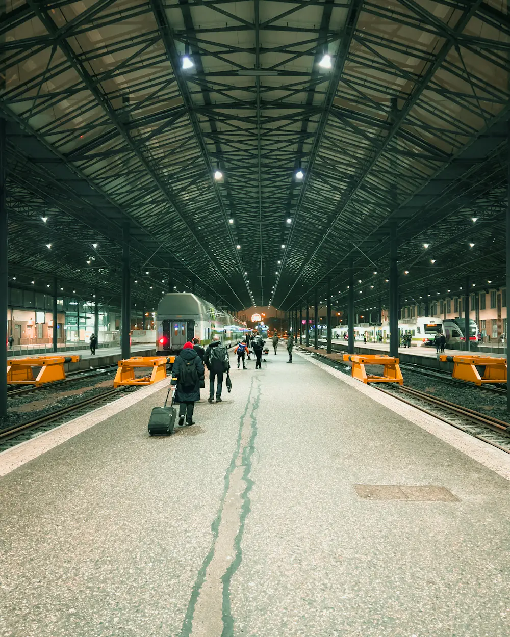 Evening view down a covered platform at Helsinki Central station, with travellers walking away from the camera towards a waiting commuter train